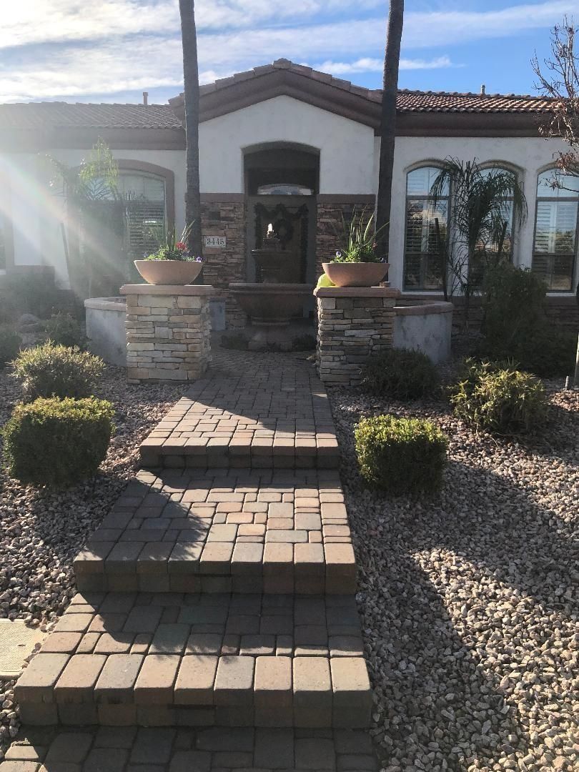 A brick pathway leads to a house with an arched doorway, steps, and potted plants.