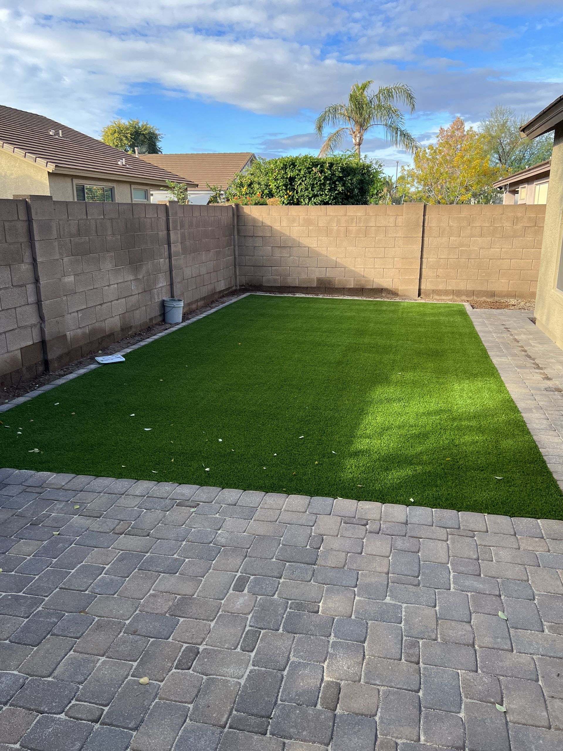 Backyard with green turf surrounded by a stone patio and beige walls under a cloudy sky.