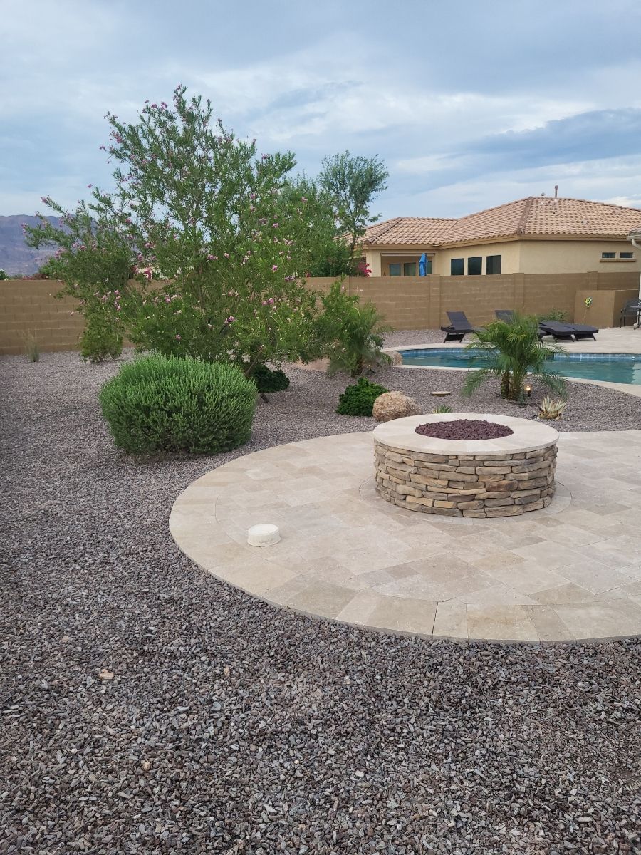 Backyard with a stone fire pit, pool, and desert landscaping under a cloudy sky.
