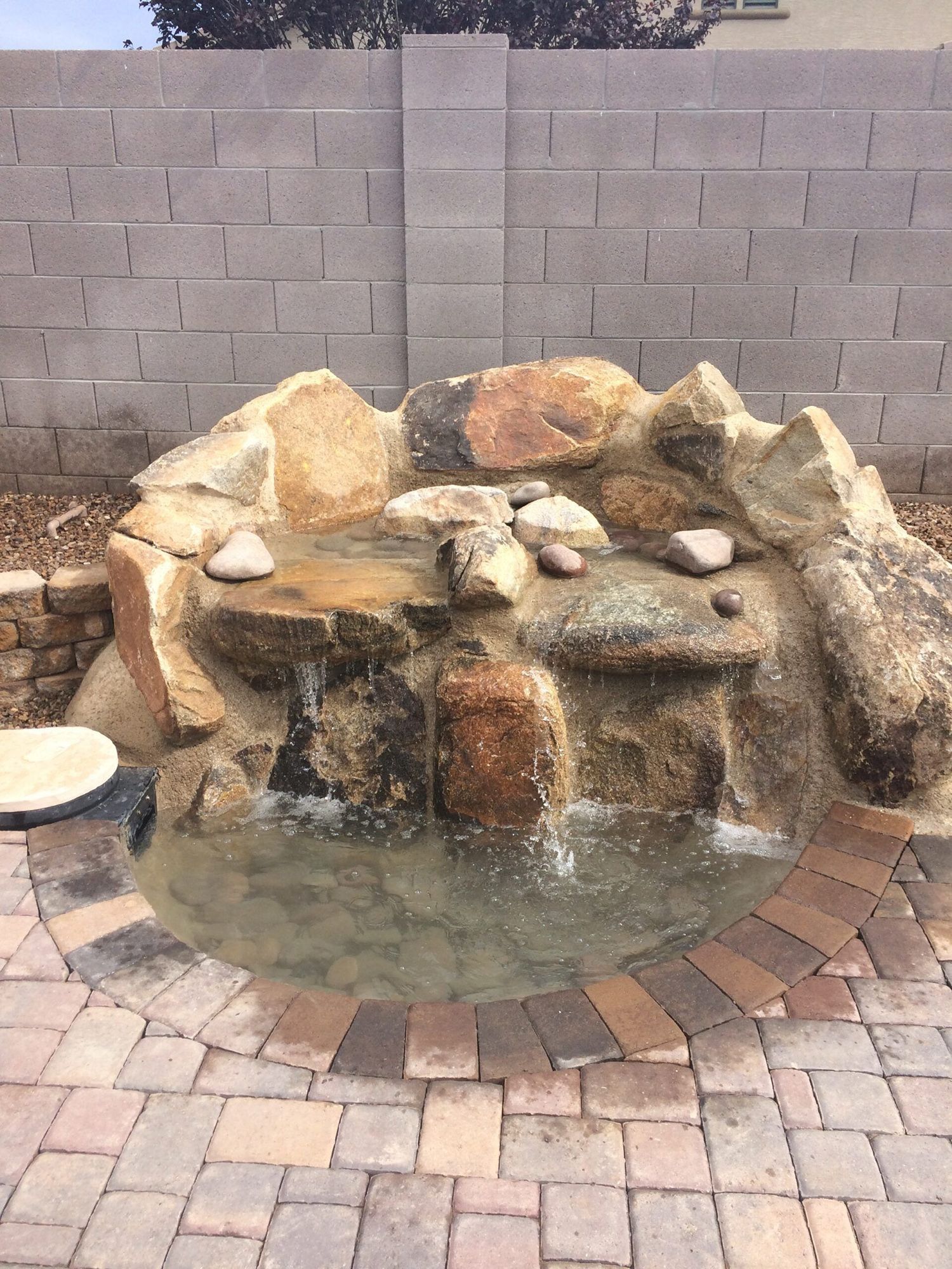 Stone waterfall feature with water flowing into a small pond, surrounded by brick pavers, against a gray block wall.