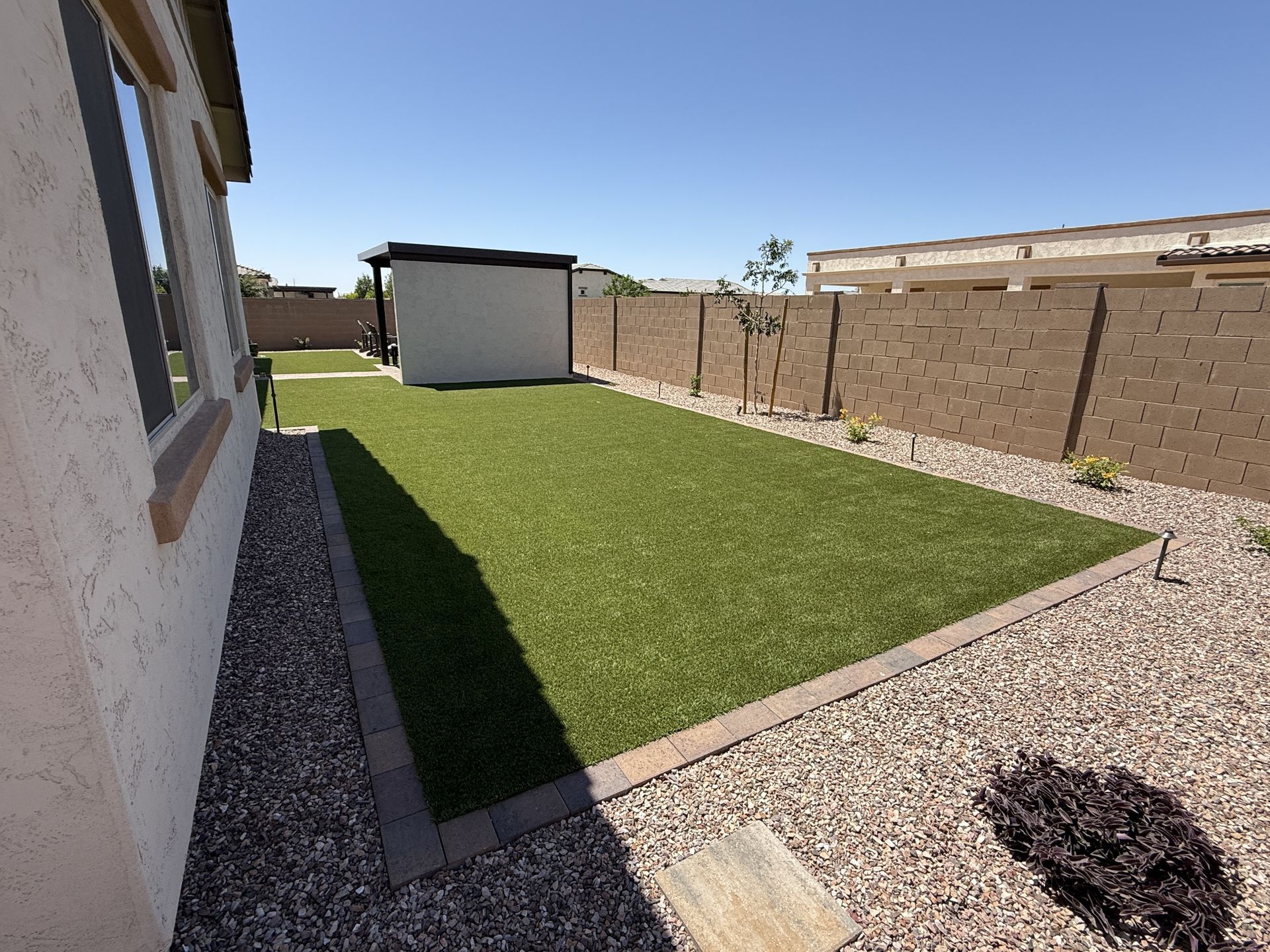 Backyard with green artificial turf, rock border, tan stucco walls, and a small shed under a blue sky.