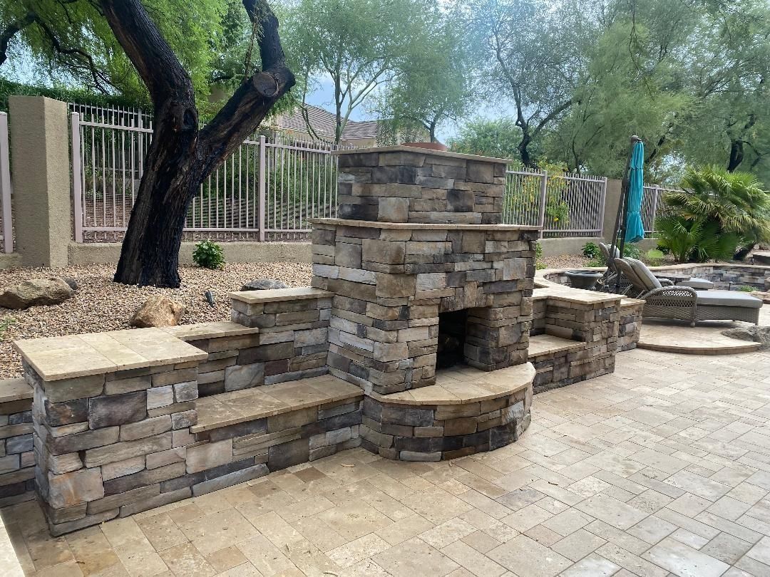 Stone outdoor fireplace with built-in seating on a paved patio, fence and trees in background.