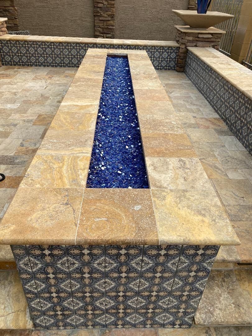 Long rectangular fire pit with blue glass, surrounded by stone and patterned tiles.