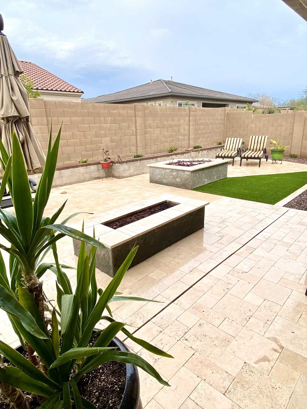 Patio with two fire pits, lawn, and chairs against a beige wall, with a plant in the foreground.