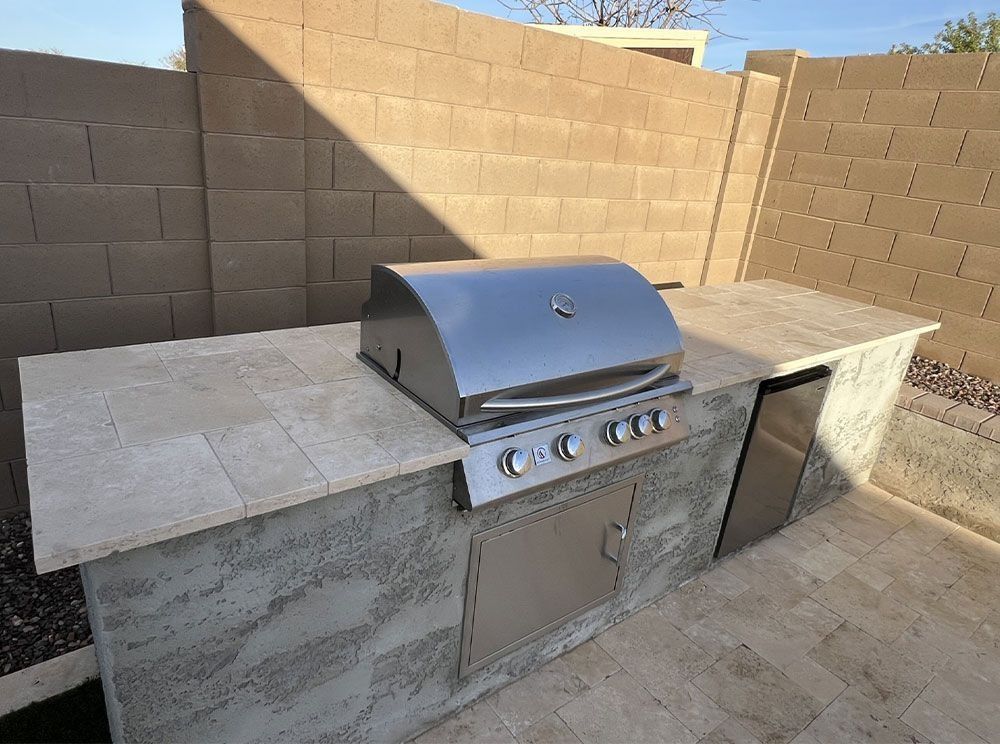 Outdoor built-in kitchen with stainless steel grill and countertop, beige walls, and concrete patio.