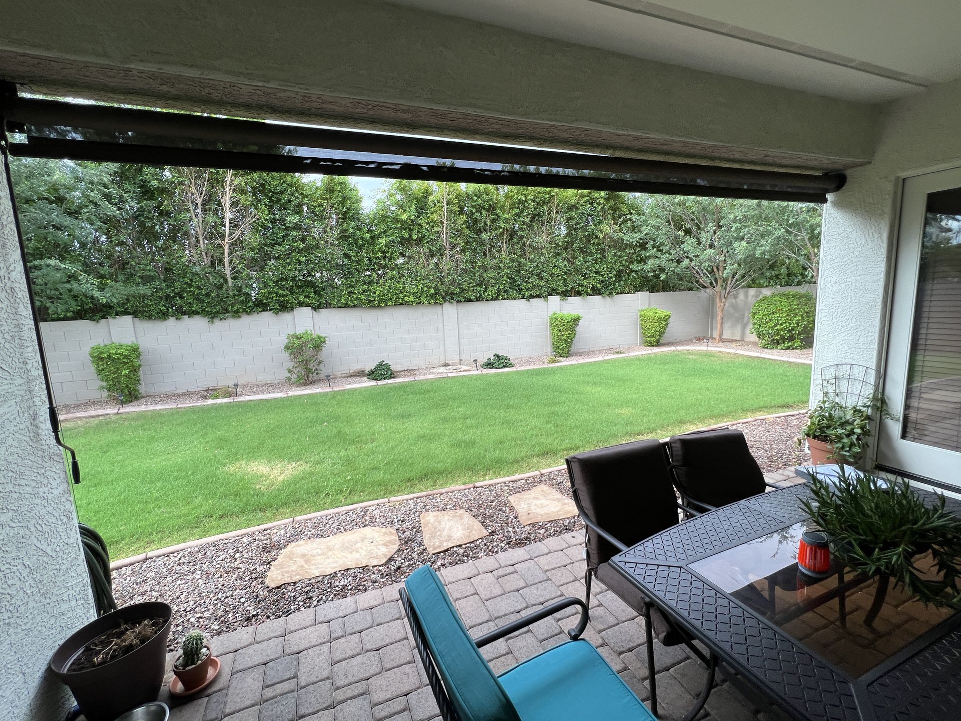Patio with black shade, table with chairs, and a backyard with green grass, bushes, and a stone border.