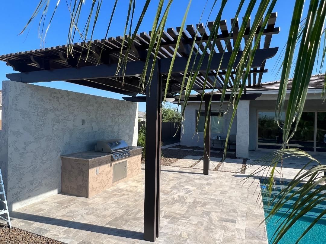 Pergola structure over a stone patio with a built-in outdoor kitchen. Pool visible in the background.