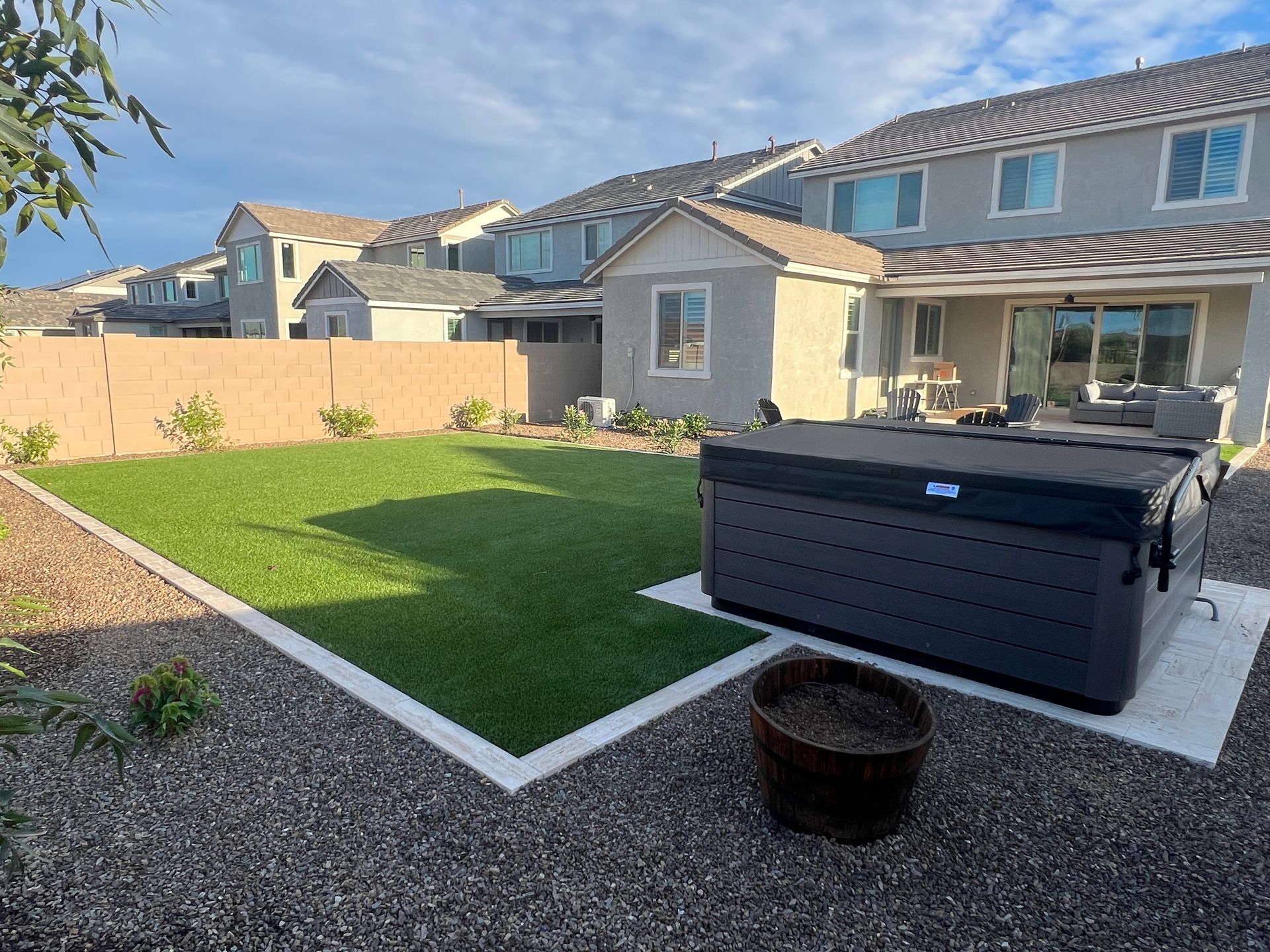 Backyard with hot tub on concrete pad, green turf lawn, gravel, beige fence and houses in the background.