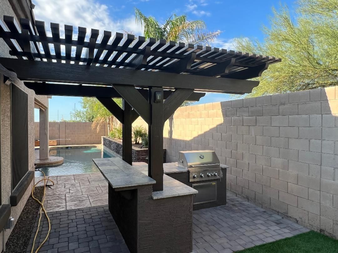 Outdoor kitchen and bar area with a grill, pool, and pergola under a blue sky.