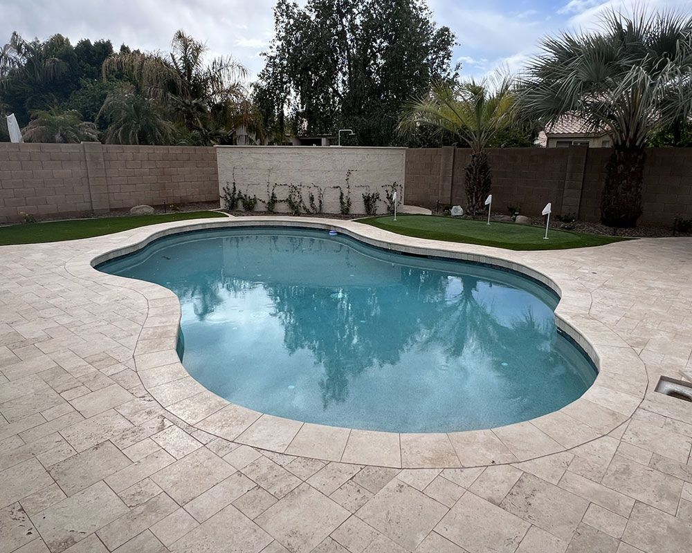 Swimming pool with turquoise water, surrounded by light-colored stone patio and grass, walled in yard.