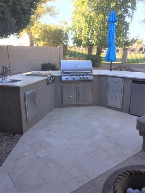 Outdoor kitchen with stainless steel grill, sink, and cabinets on a tiled patio. Blue umbrella provides shade.