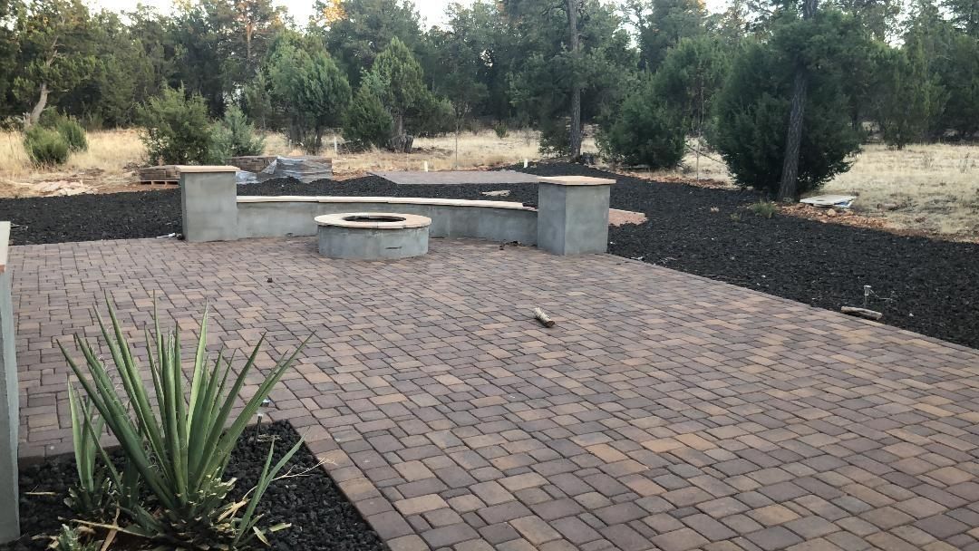 Brick patio with fire pit, built-in concrete bench, and black lava rock border, surrounded by trees and dry grass.