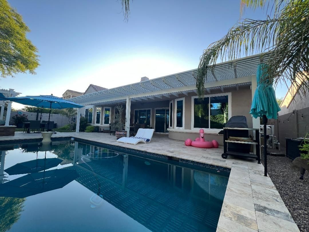 Backyard pool with patio, lounge chair, grill, and pink inflatable flamingo; bright, sunny day.