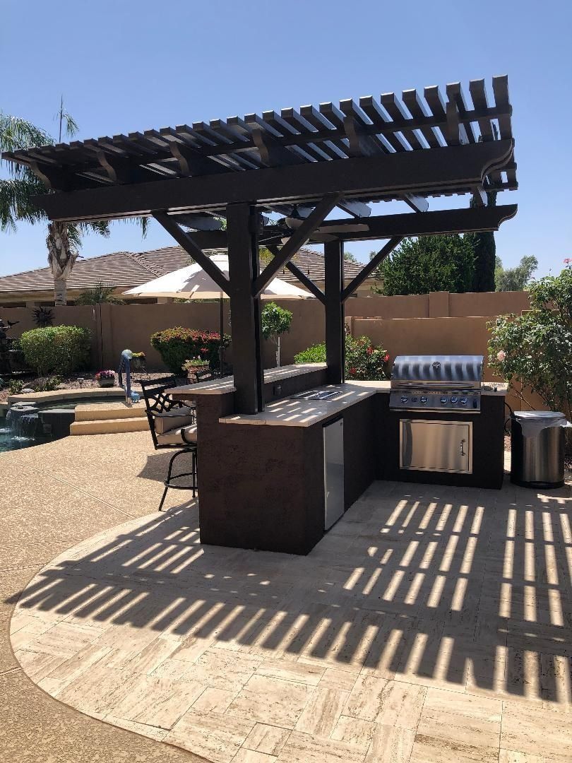 Outdoor kitchen with pergola, built-in grill, and bar seating on a sunny patio next to a pool.