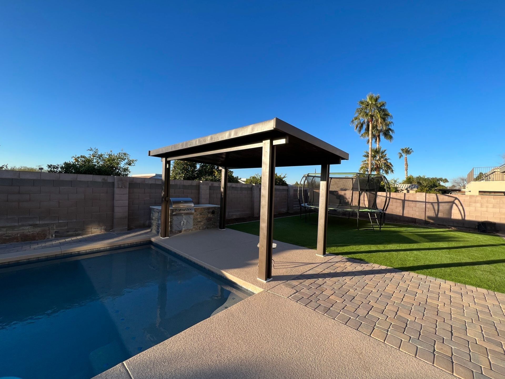 Backyard scene with a pool, grill, pergola, trampoline, and palm trees under a blue sky.