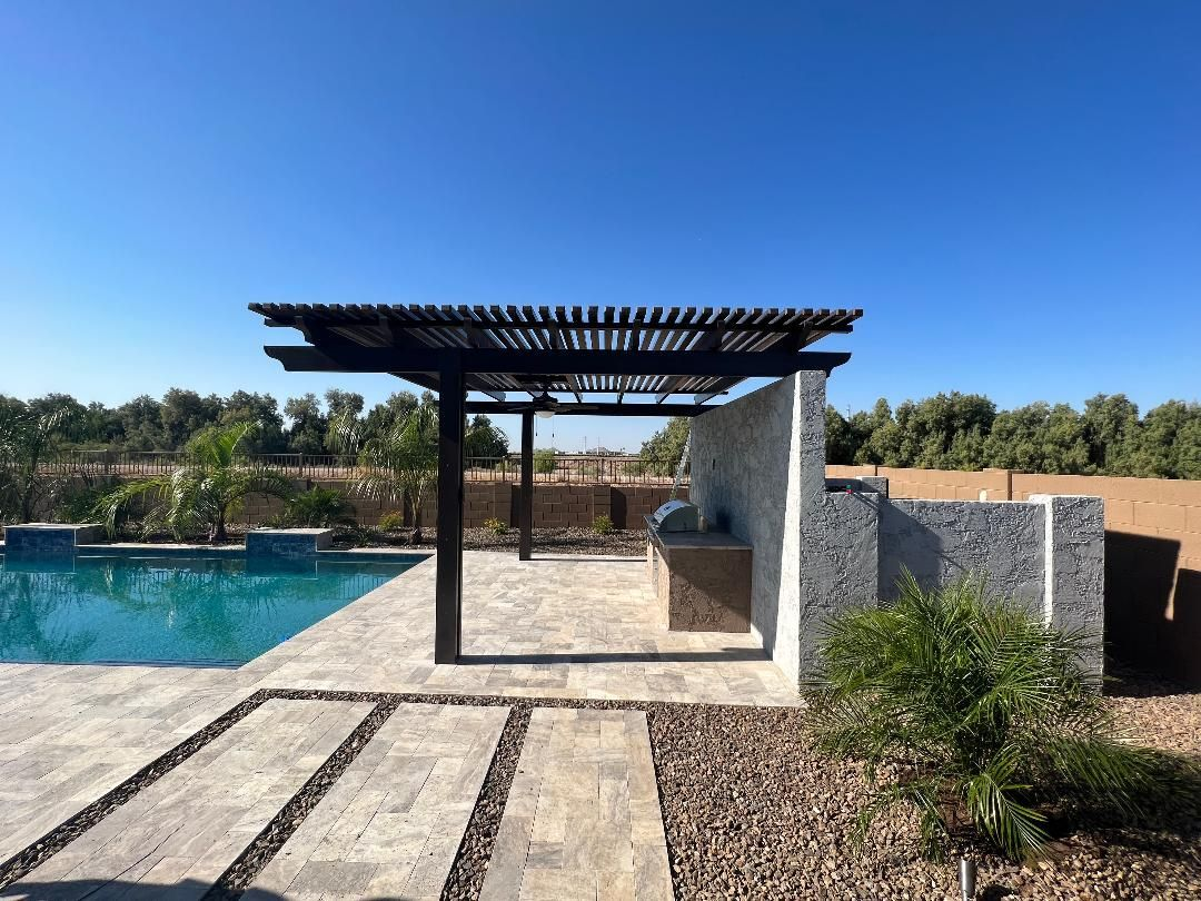 Pergola over outdoor kitchen next to a pool. Black pergola, blue sky, stone patio, and stone wall.