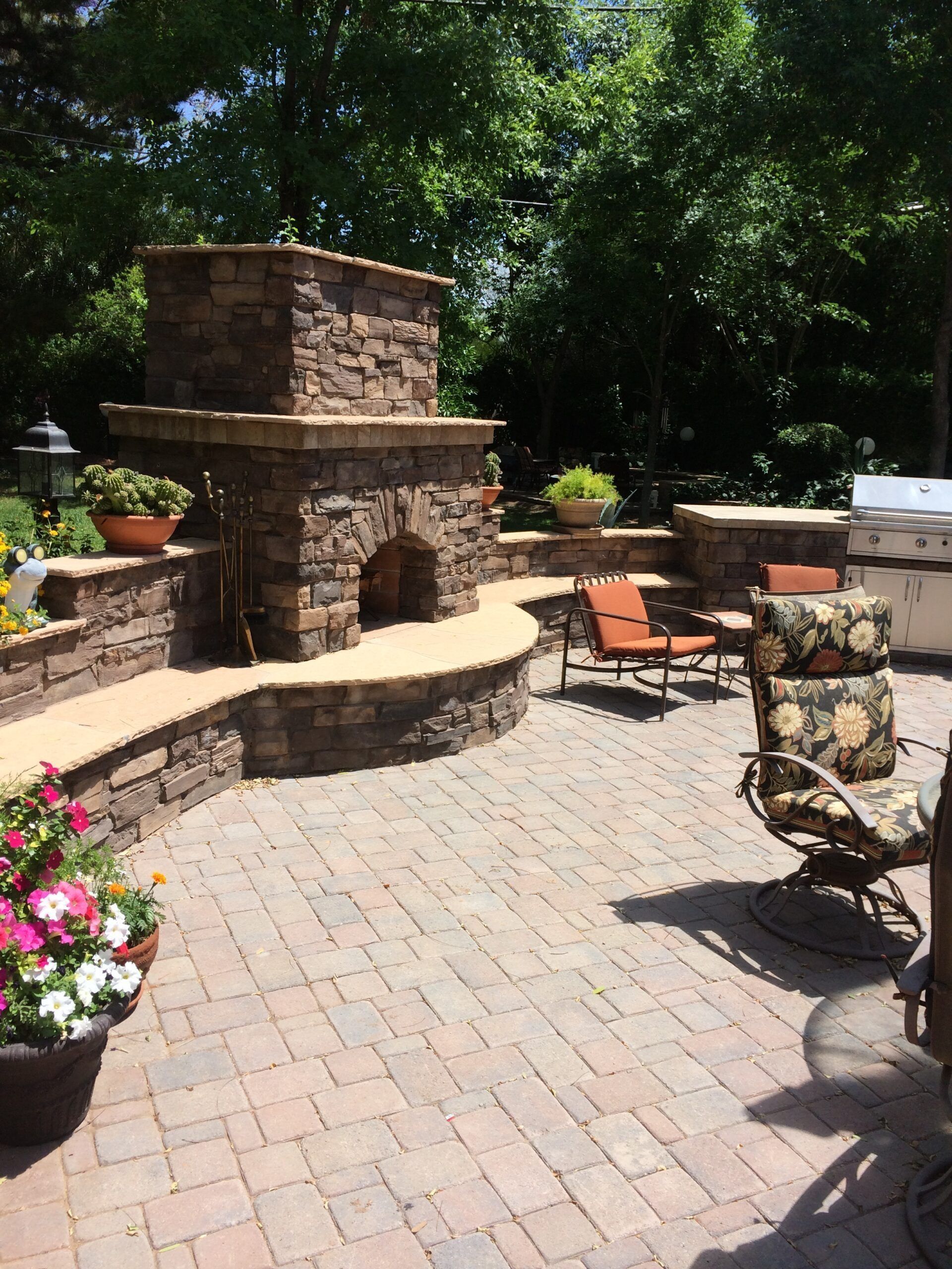 Stone fireplace with seating area, brick patio, and potted plants in an outdoor setting.