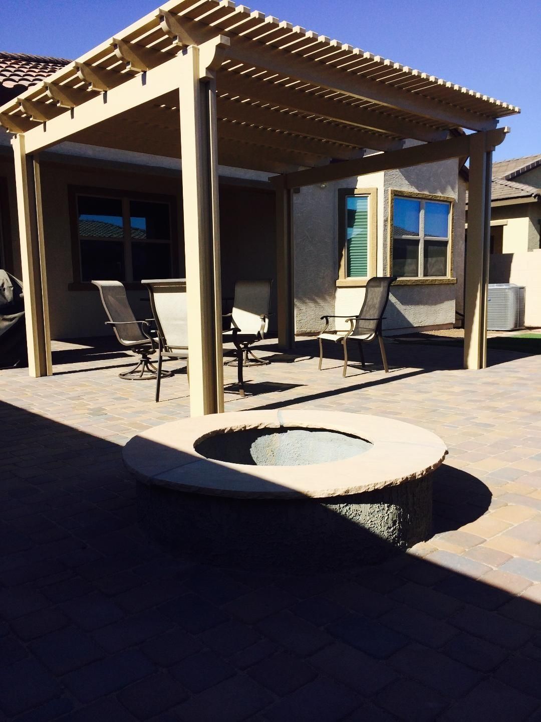 Patio with pergola, fire pit, and chairs. Beige structure on brick patio in sunlight.
