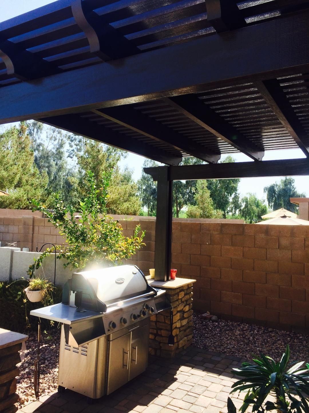 Outdoor grilling area with a stainless steel grill under a wooden pergola, set against a brick wall.