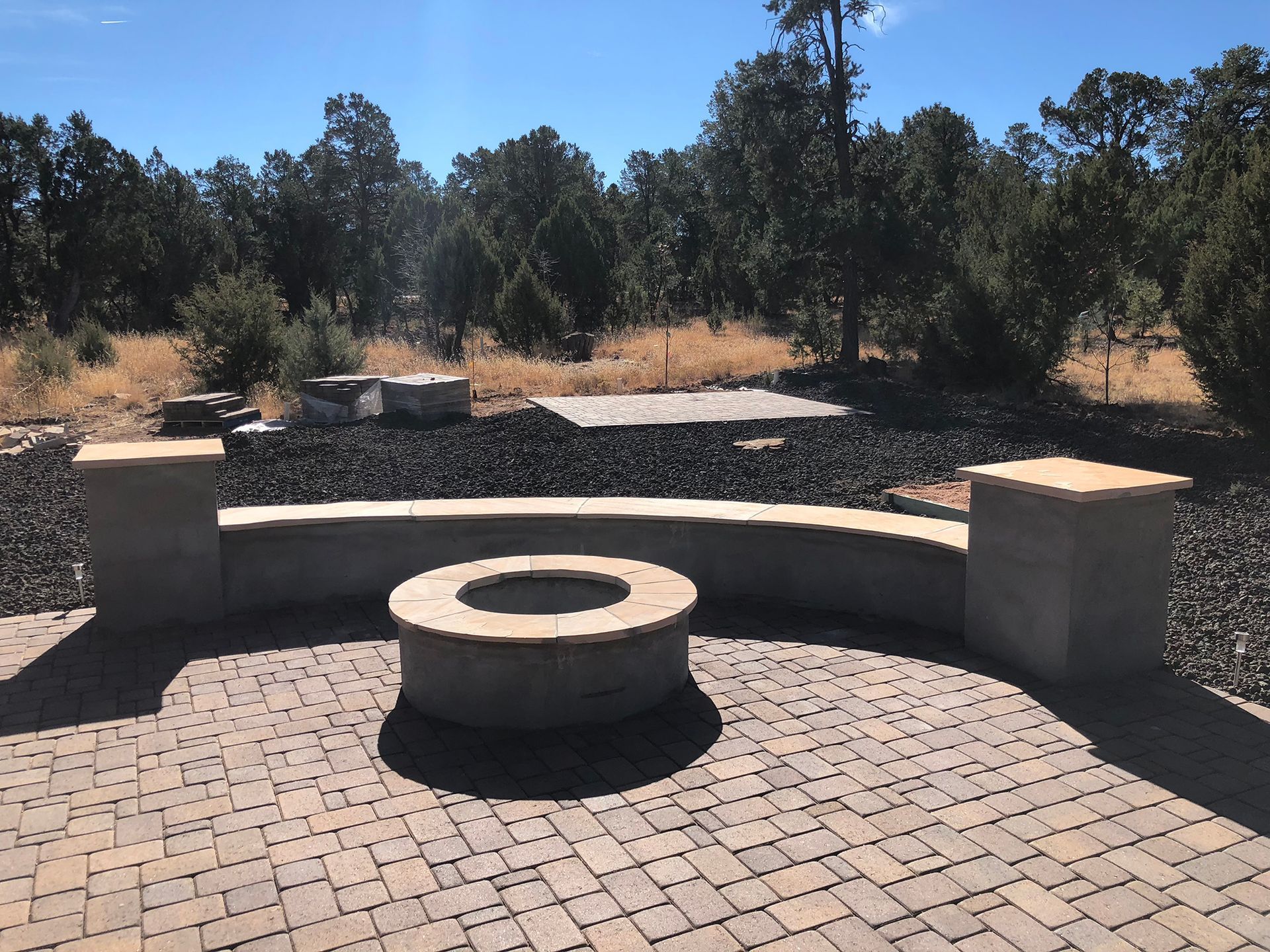 A brick patio with a circular fire pit and curved seating area, surrounded by trees.