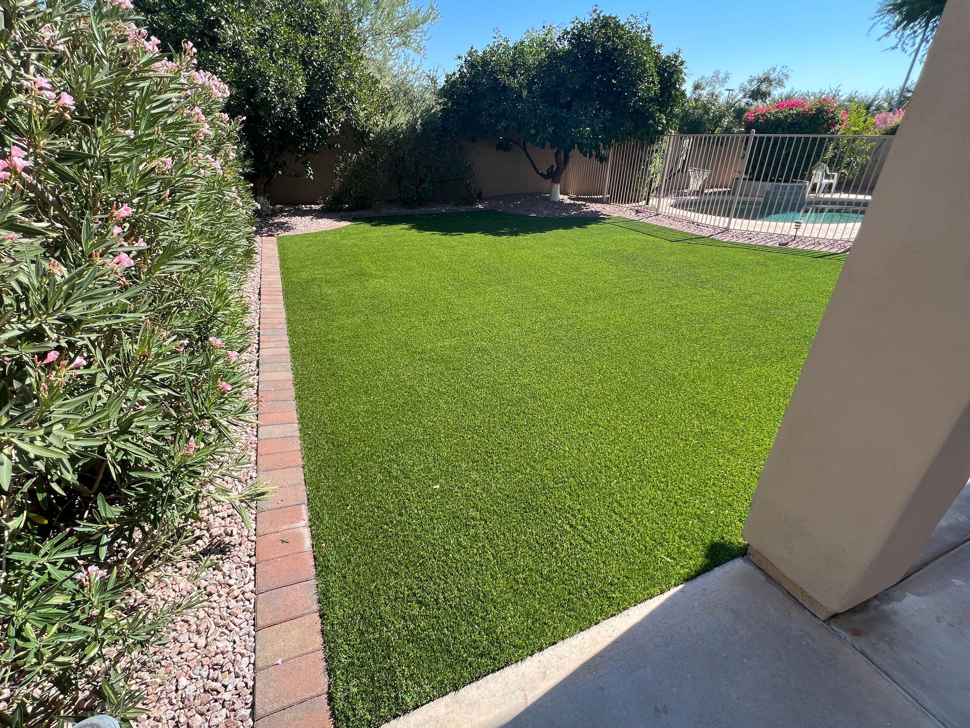 Green artificial turf in a backyard, bordered by red brick and bushes, with a concrete patio visible.
