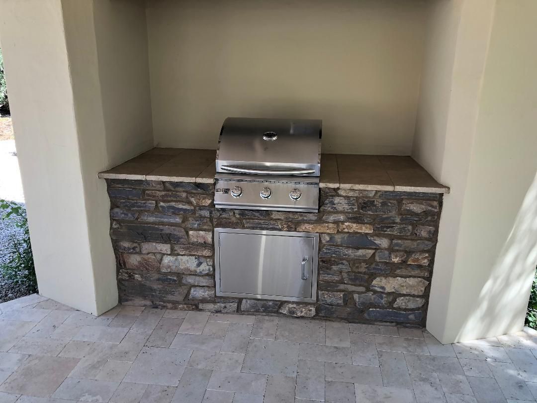 Built-in outdoor grill with stone facade, stainless steel appliances, and beige walls on a paved patio.
