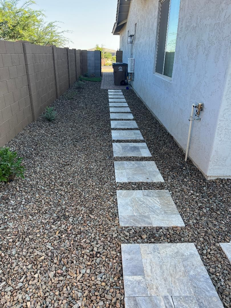 Stone path alongside a stucco house and gravel yard, leading to a trash can near a wooden fence.
