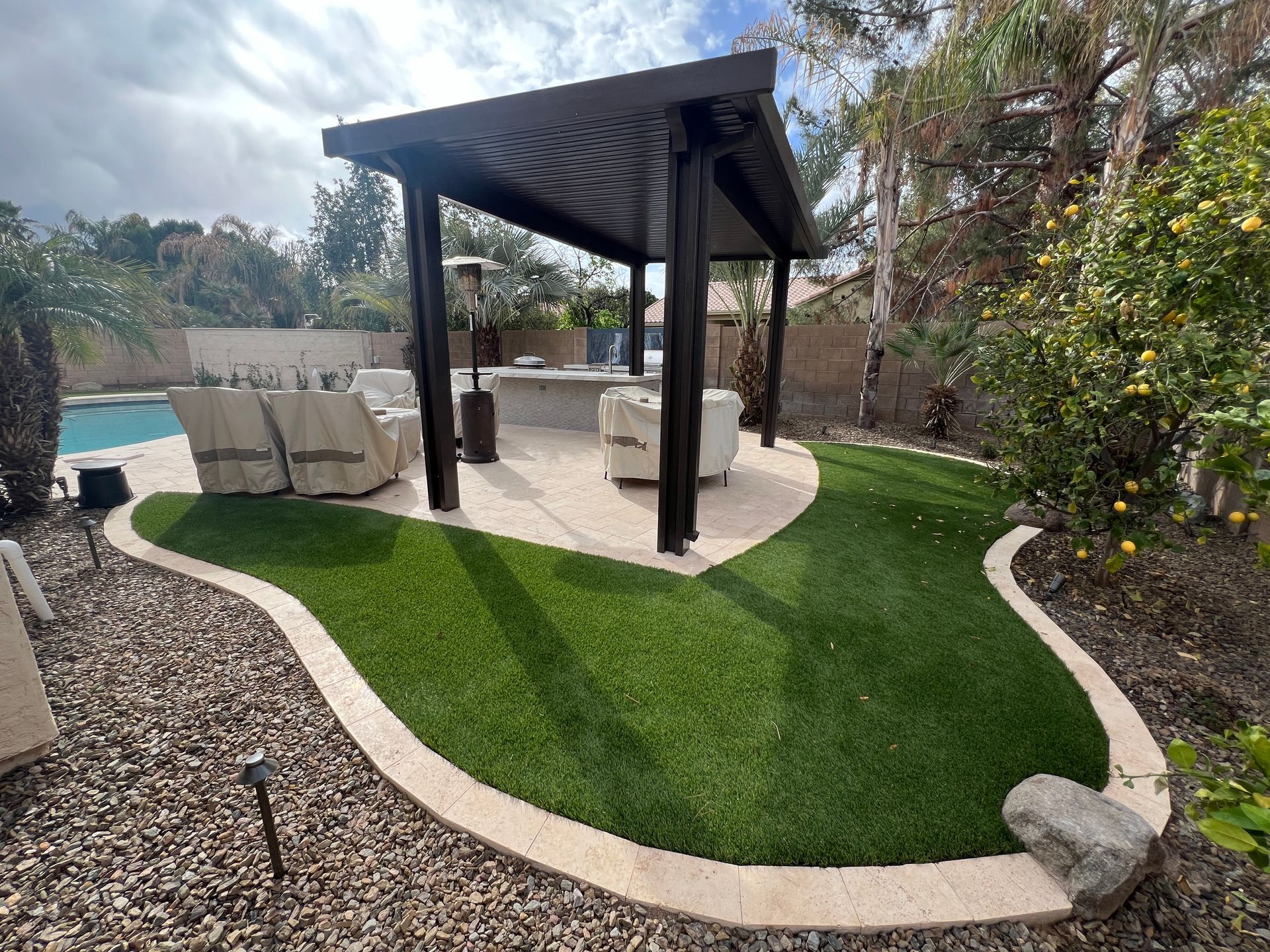 Backyard with a dark pergola over a bar, seating, and artificial grass bordered by a concrete walkway.
