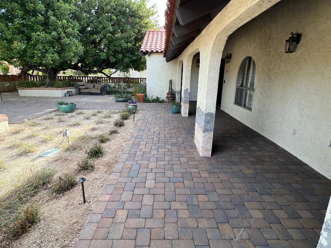 Brick walkway alongside a white stucco building with arched supports, green plants, and a tree.