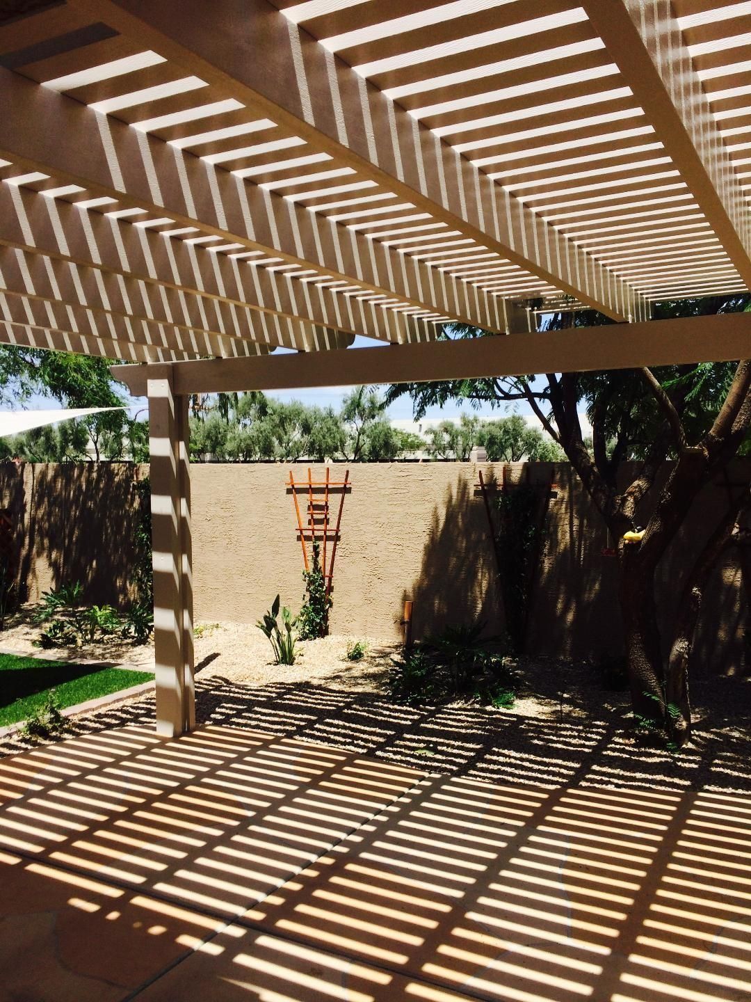 Pergola with shadows over patio and wall, desert landscaping in background.