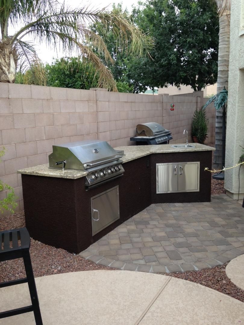 Outdoor kitchen with two grills, built-in cabinets, and a stone countertop against a concrete block wall.