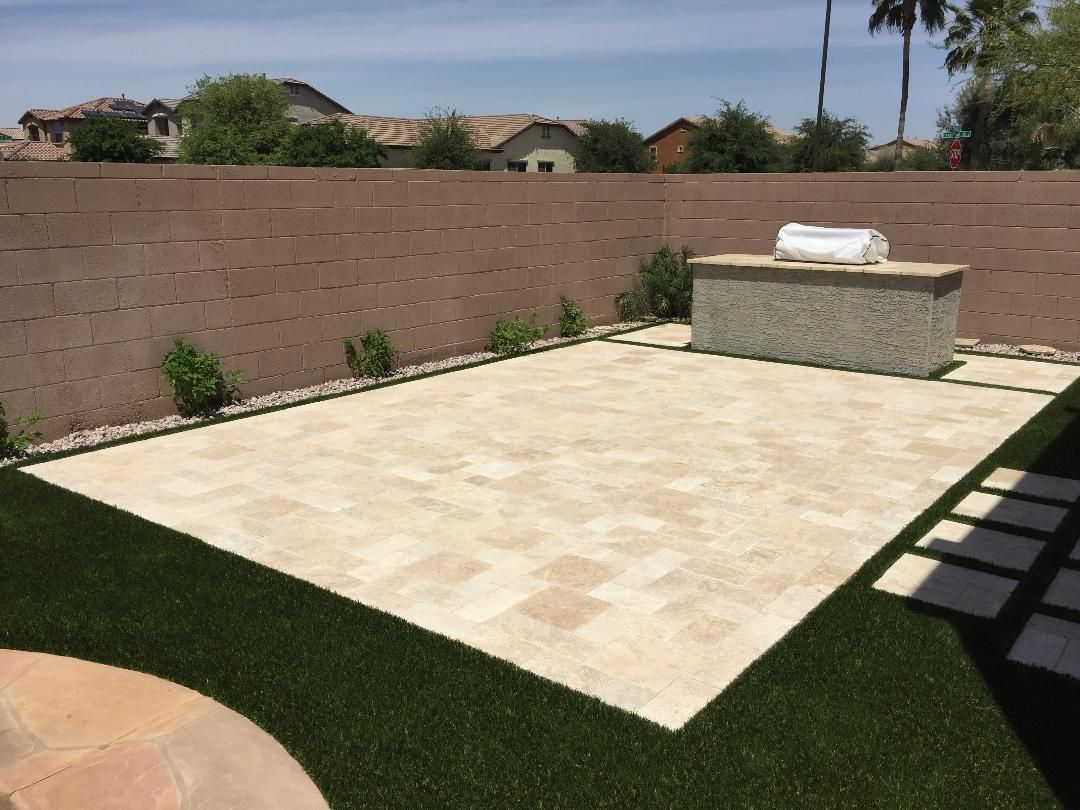 Backyard patio with light pavers, grill, and artificial turf, surrounded by a brown wall, under a sunny sky.