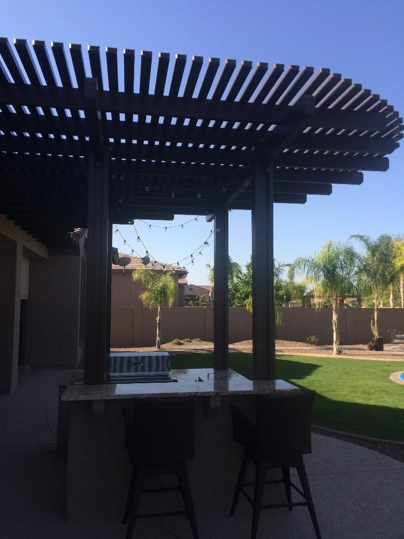 Outdoor bar with dark brown pergola and stools, string lights, and green lawn in backyard.