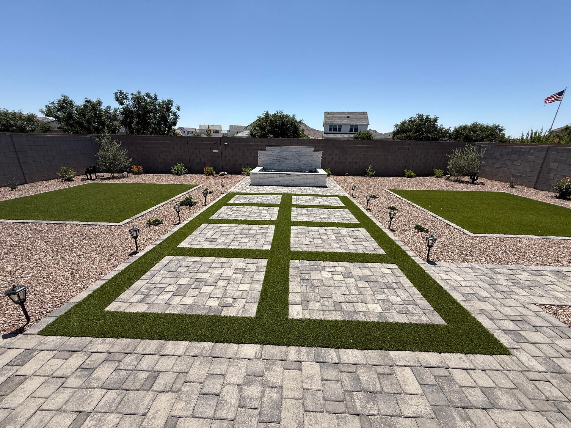 Landscaped backyard with stone patio, turf squares, and fountain against a blue sky.