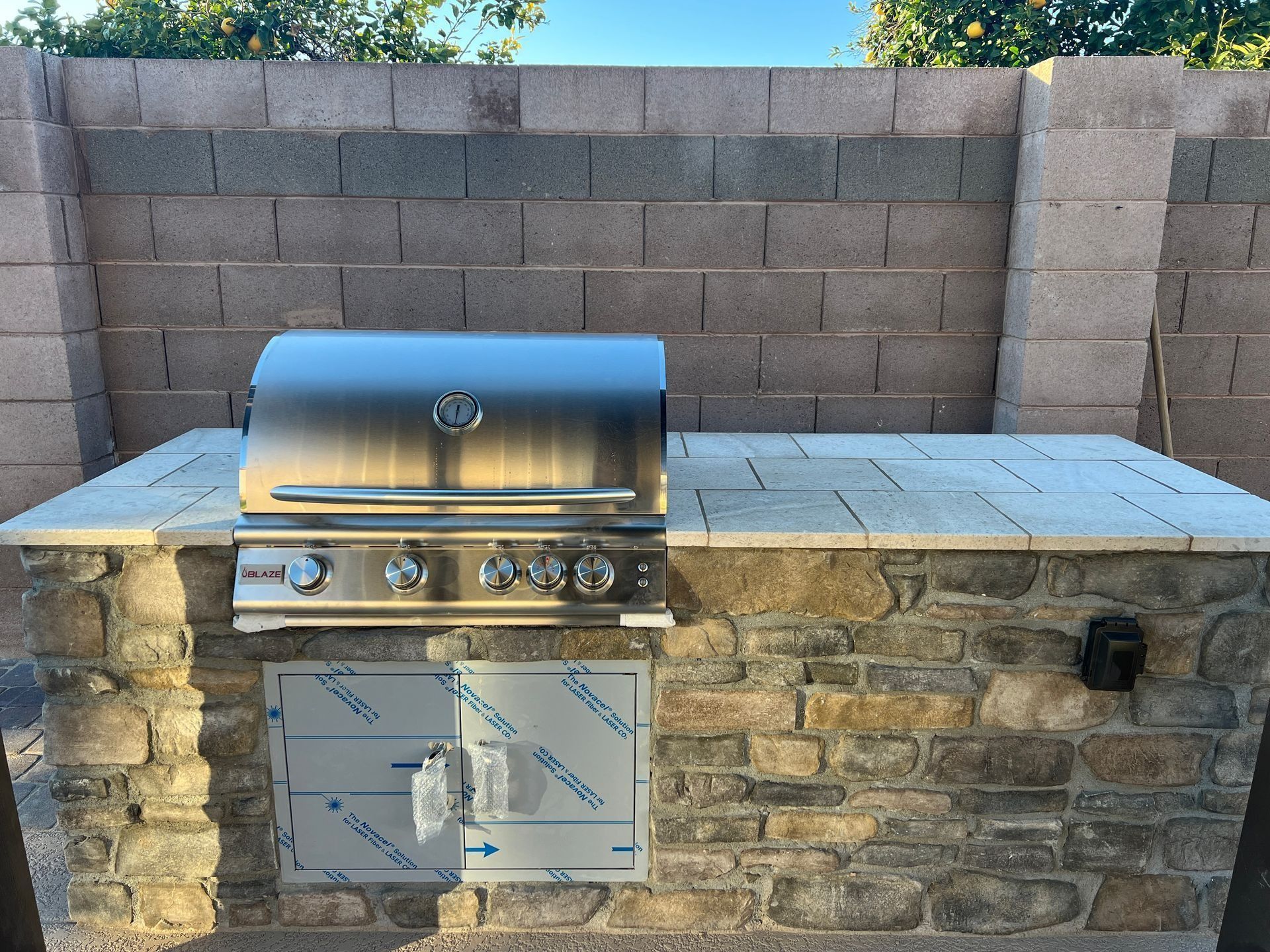Built-in outdoor kitchen with stainless steel grill and stone facade against a cinder block wall.