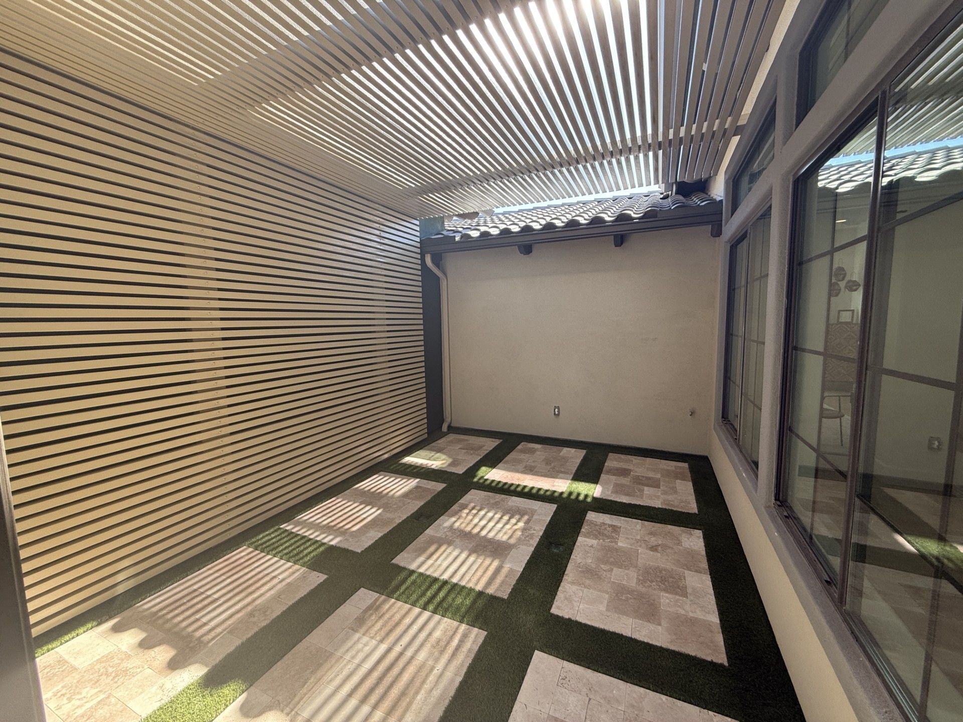 Outdoor patio with a slatted roof, tiled floor, and artificial grass. Sunlight creates shadows.