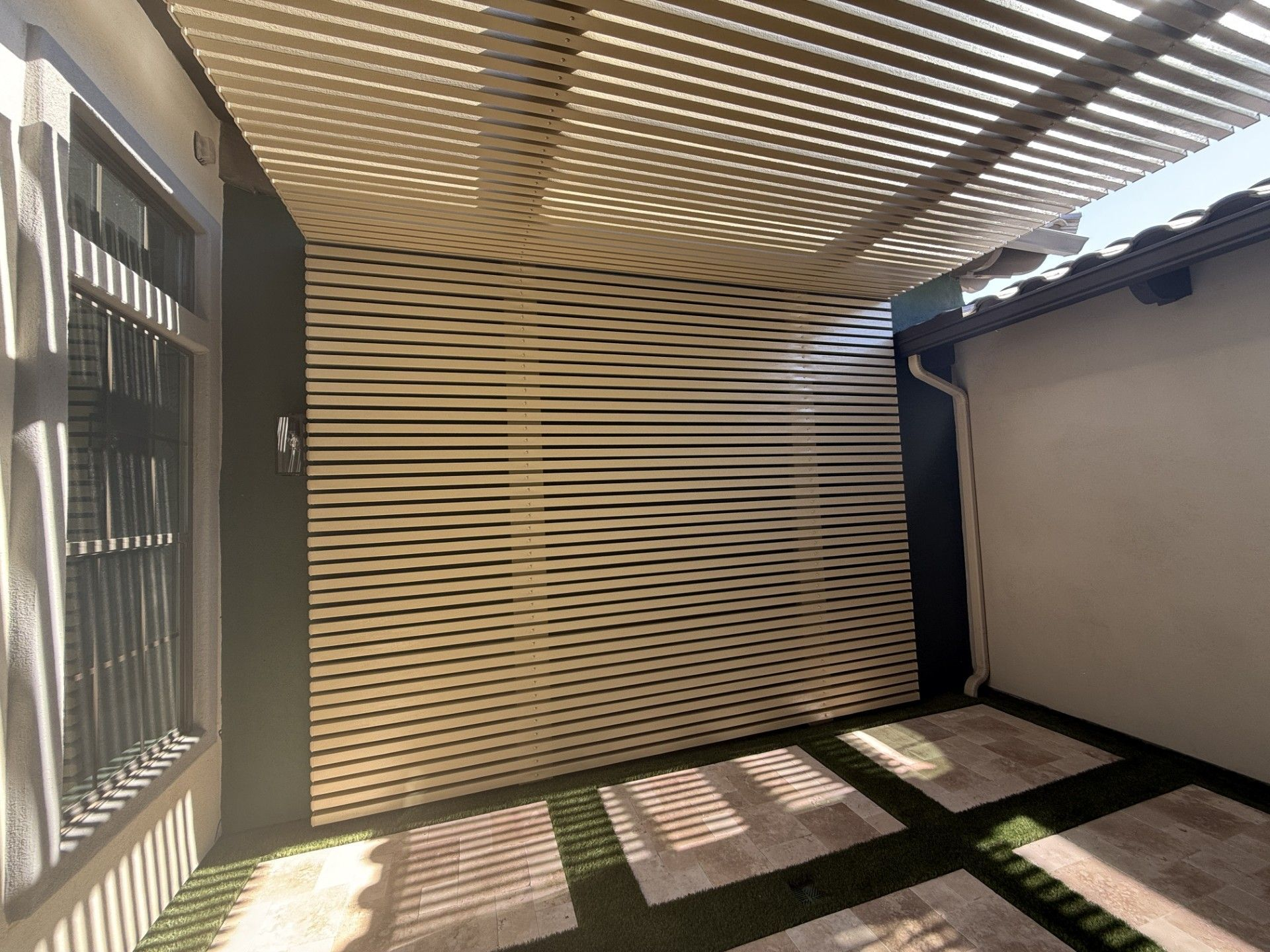 Covered outdoor patio with a slatted roof, stone flooring, and a horizontal wood slat wall.