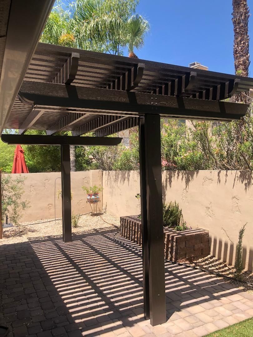 Pergola with striped shadows on a brick patio. Brown structure against a light brown wall.