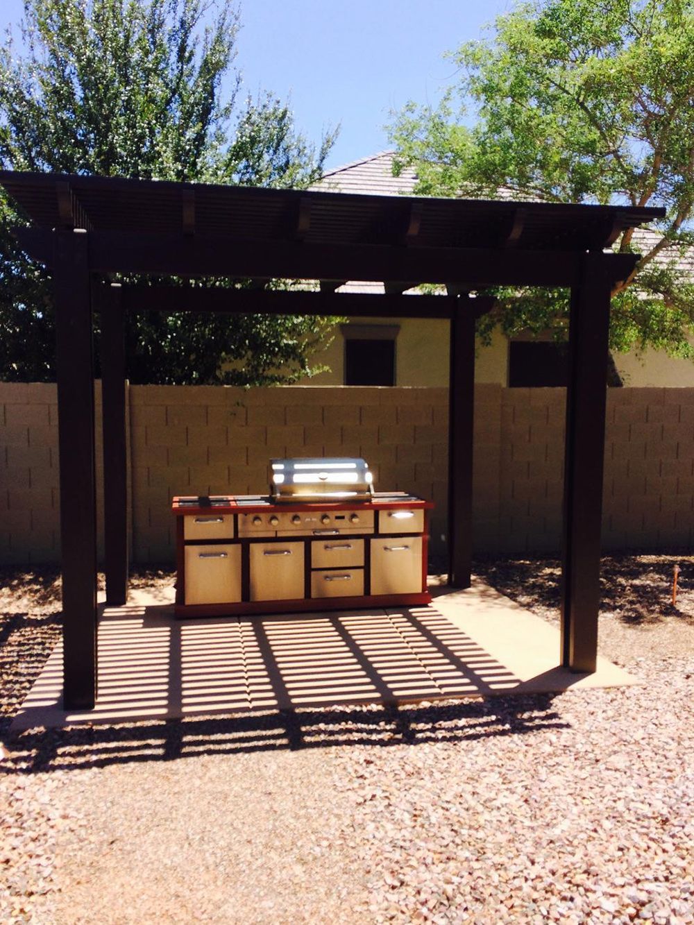 Outdoor kitchen with a grill under a dark wooden pergola, in a backyard.