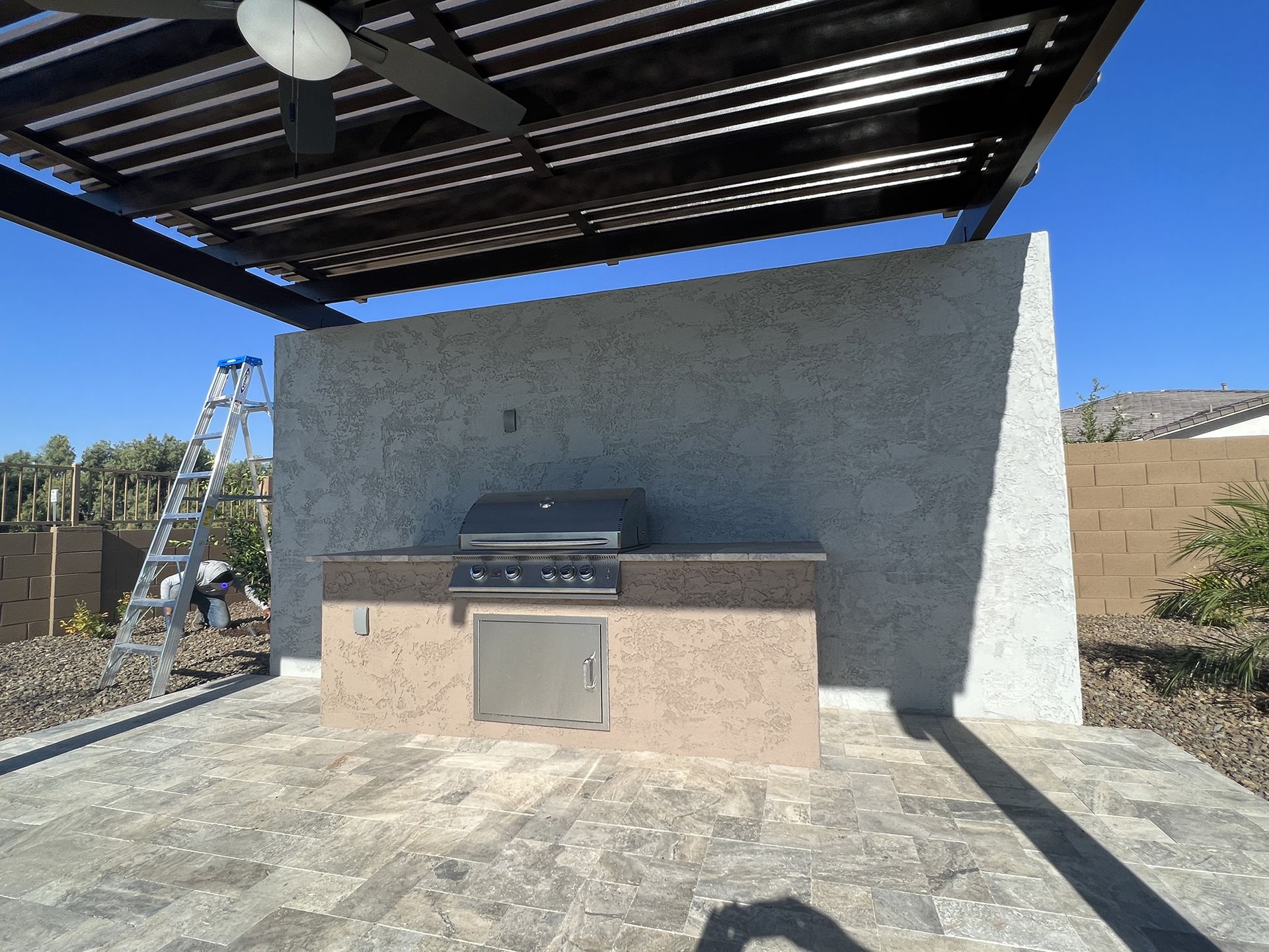 Outdoor kitchen with grill, under a pergola on a stone patio. A ladder is propped against a wall.