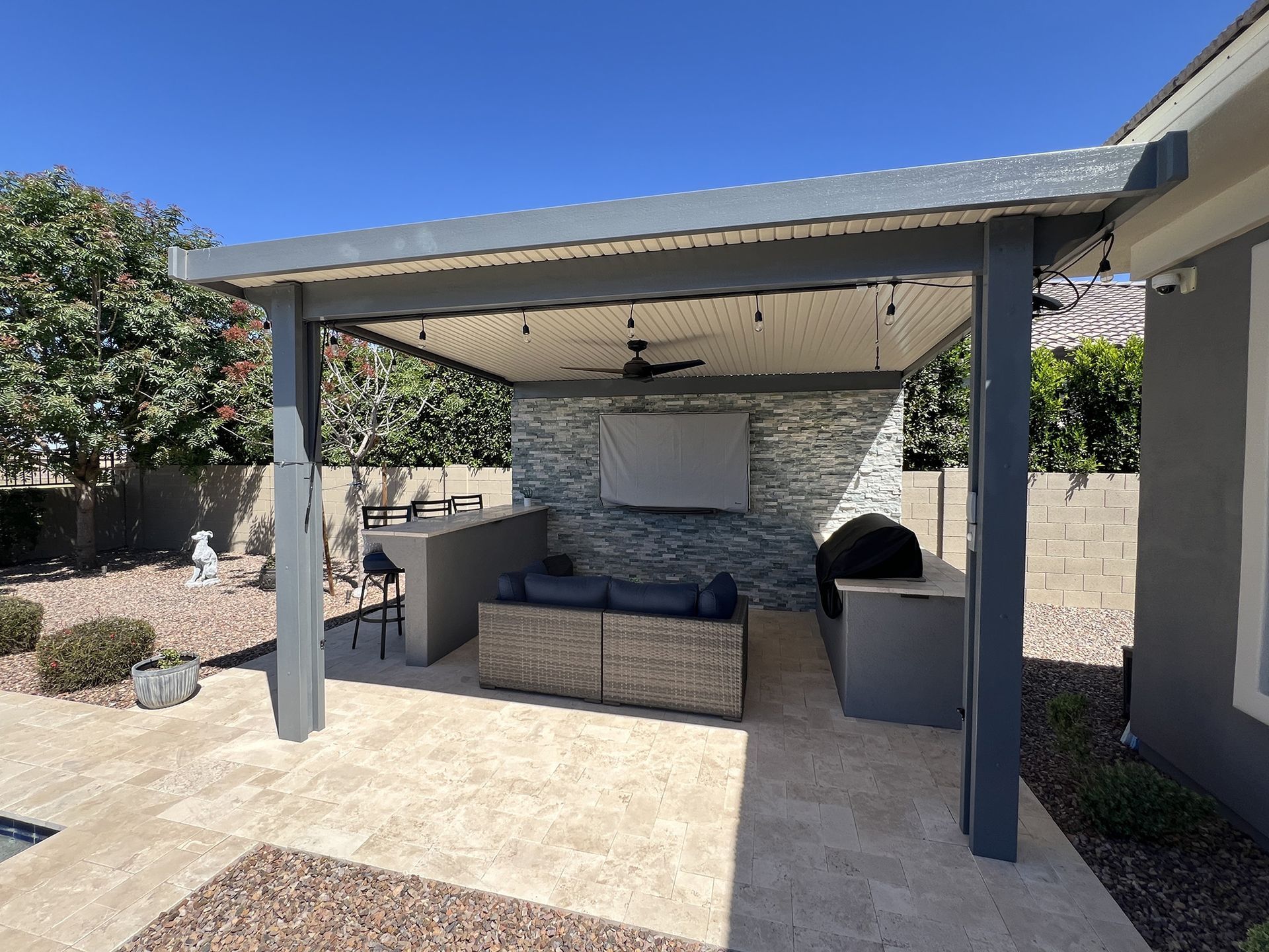 Outdoor kitchen with a stone wall, couch, grill, and TV under a pergola.