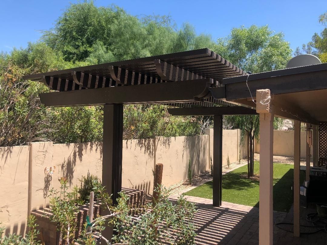 Wooden pergola with shade over a backyard patio with a wall and greenery.