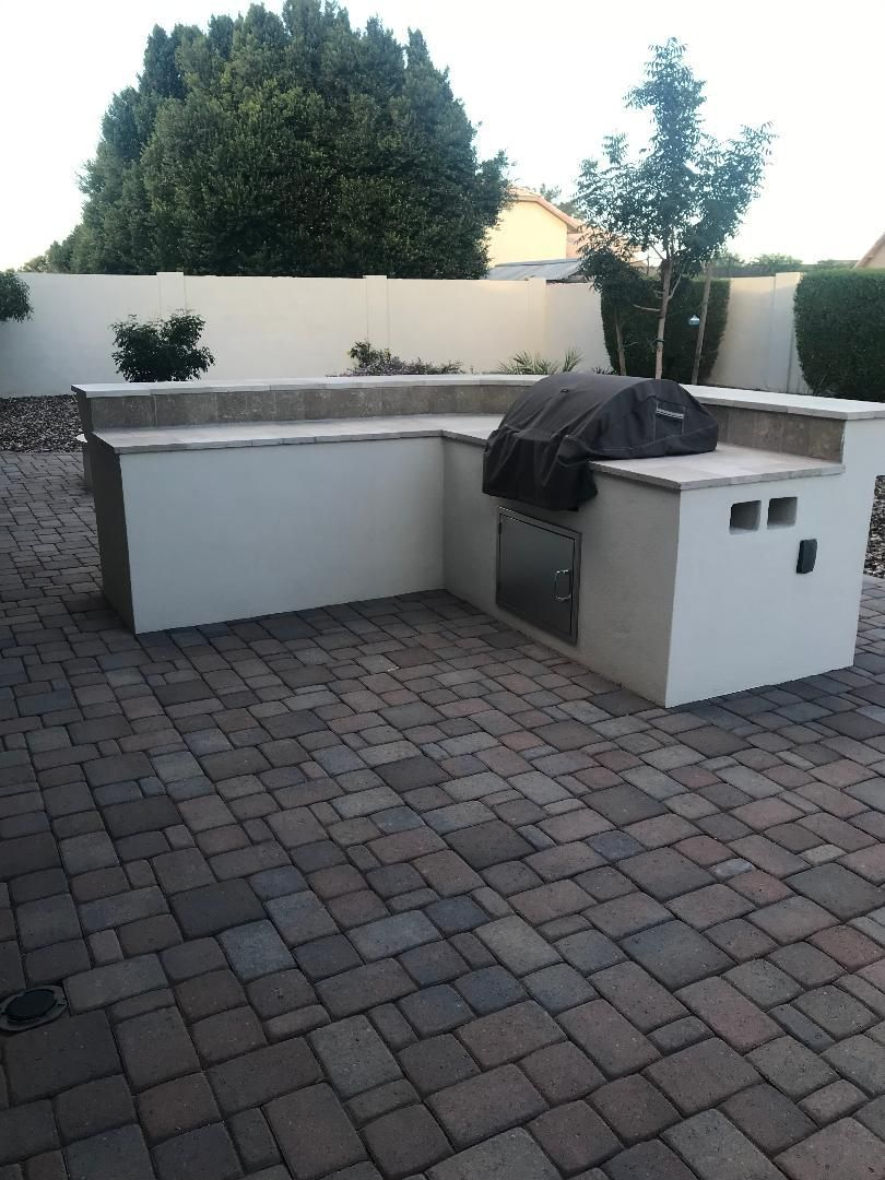 Outdoor kitchen with gray brick patio and light beige countertops. Includes a grill, storage, and small wall cutouts.