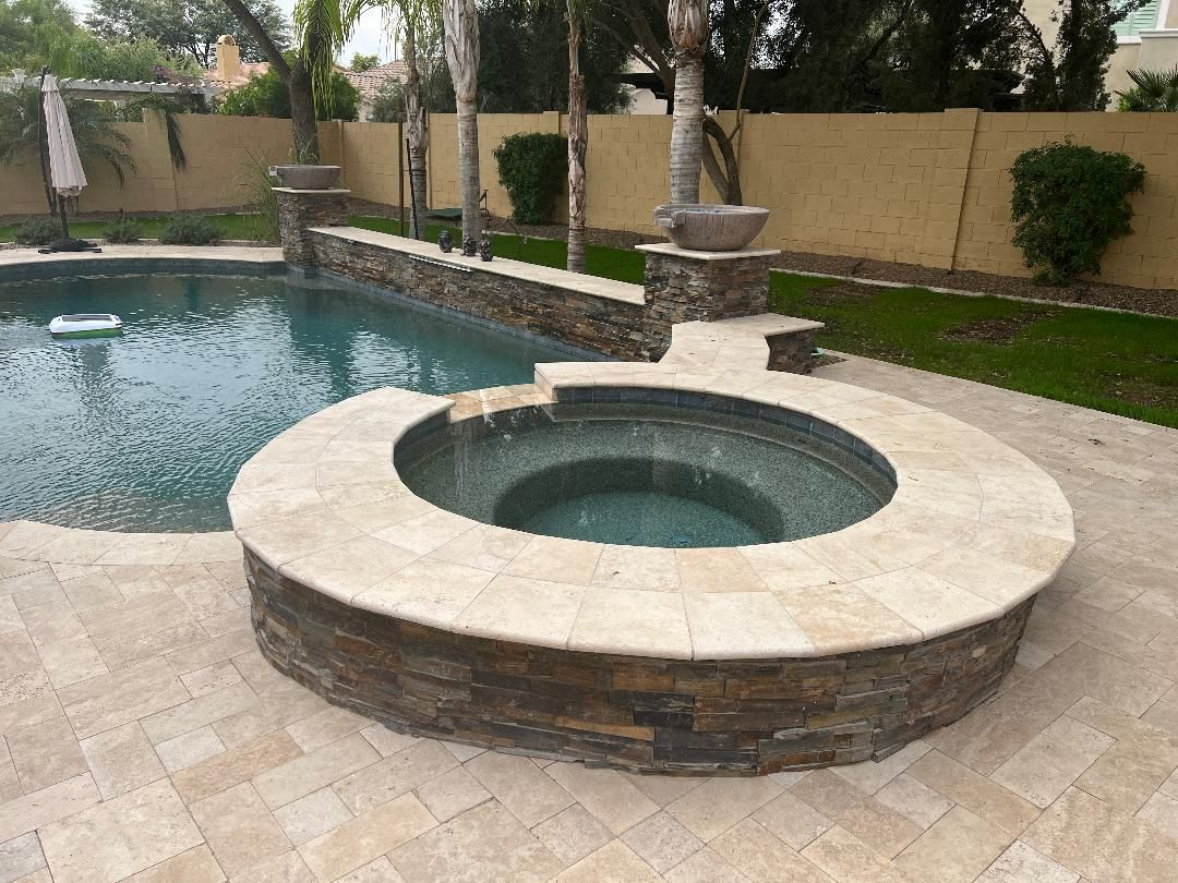 A backyard pool with a hot tub surrounded by stone pavers and a raised stone wall.