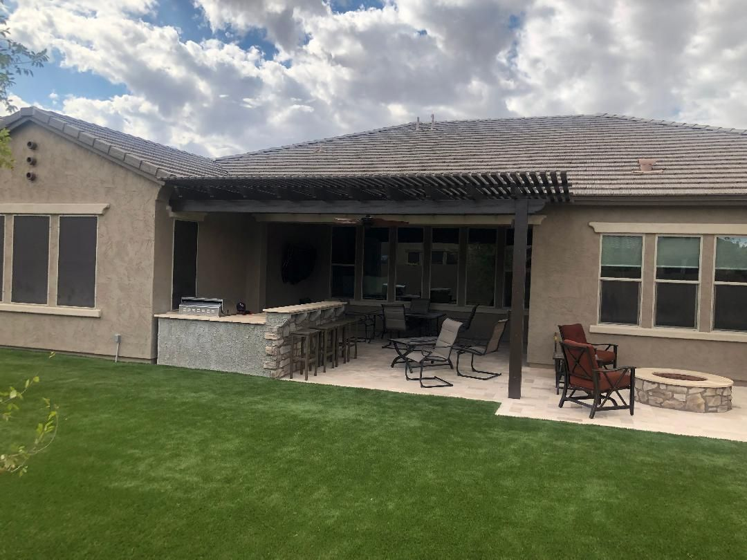 Backyard patio with a grill, seating, and fire pit under a shaded pergola, on a sunny day.