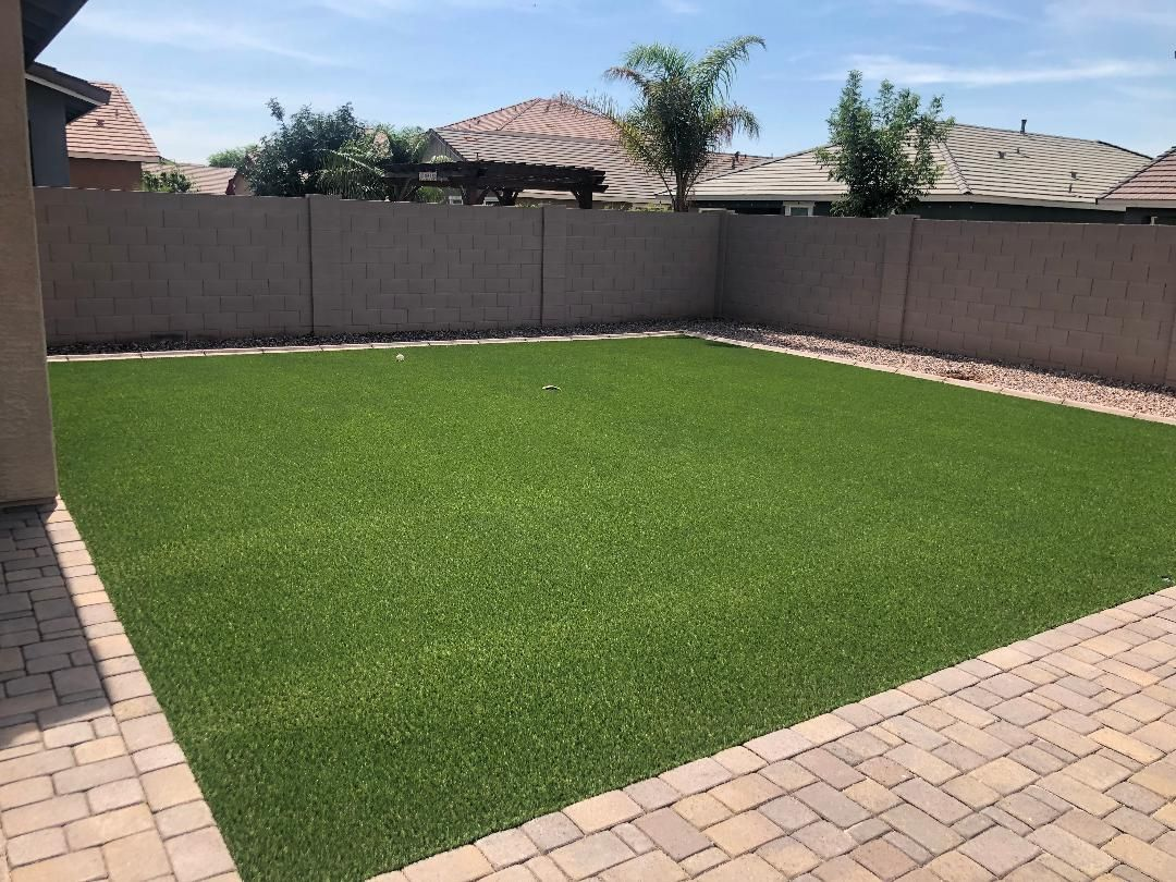 Green artificial grass in a backyard, surrounded by a stone patio and concrete walls under a blue sky.