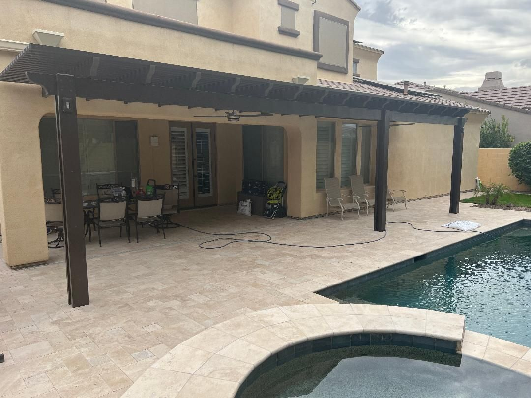 Pergola over patio next to a pool, tan stone, dark brown wood, beige house, cloudy sky.