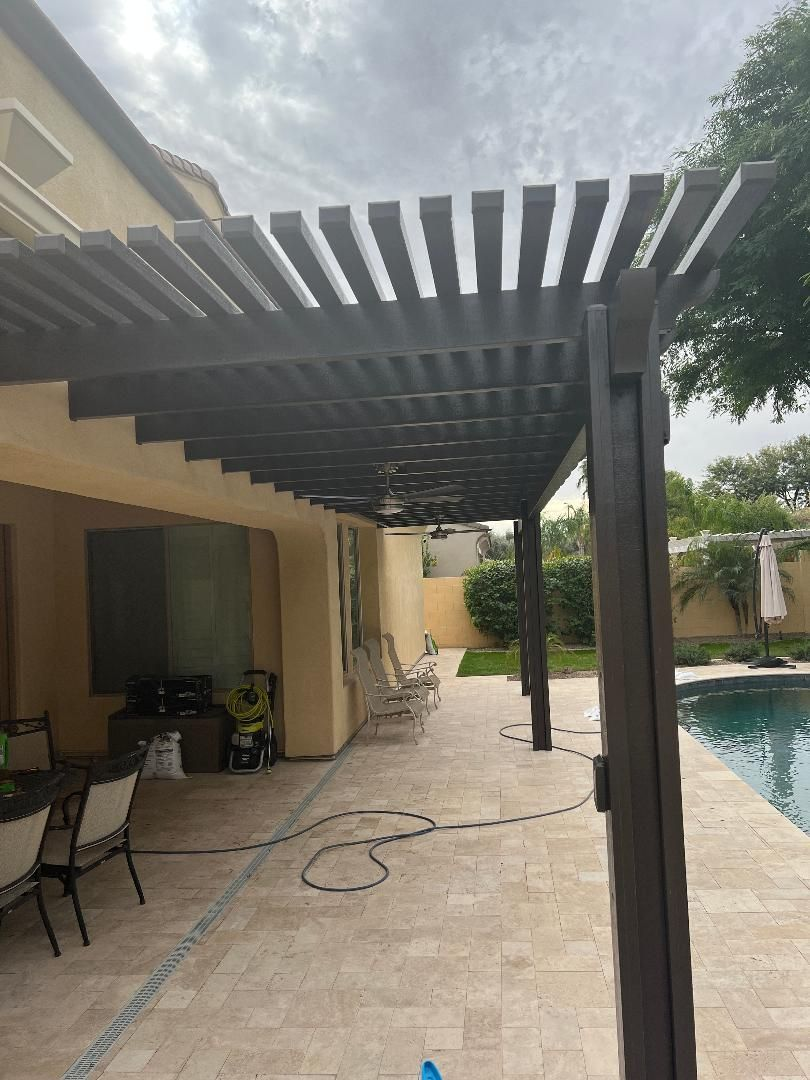 Pergola over a patio by a pool, extending from a beige house. Brown wood, cloudy sky.