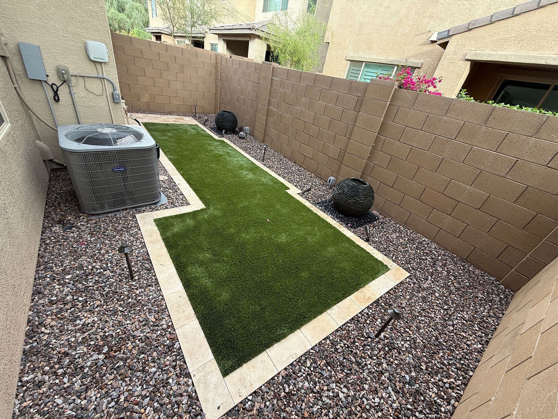 Small backyard with artificial turf, bordered by stone and beige walls, with an AC unit.