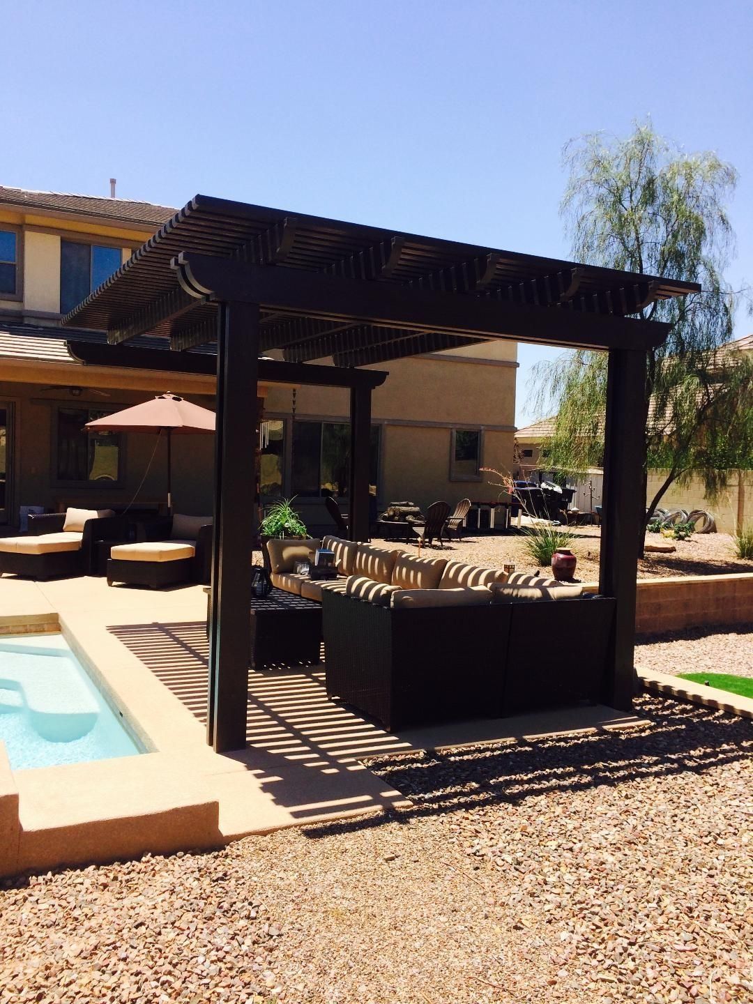 Pergola with seating by a pool in a backyard, casting shadows on the ground.